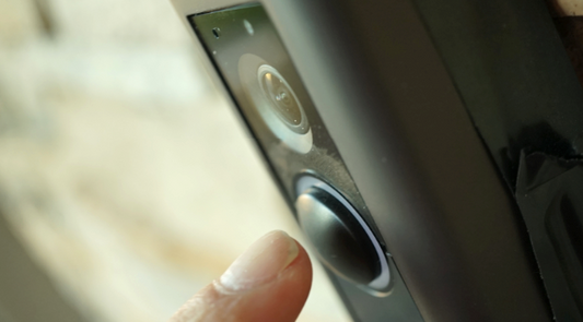 A close-up image showing a person's finger pressing the doorbell button on a modern, smart video doorbell system mounted on a wall.