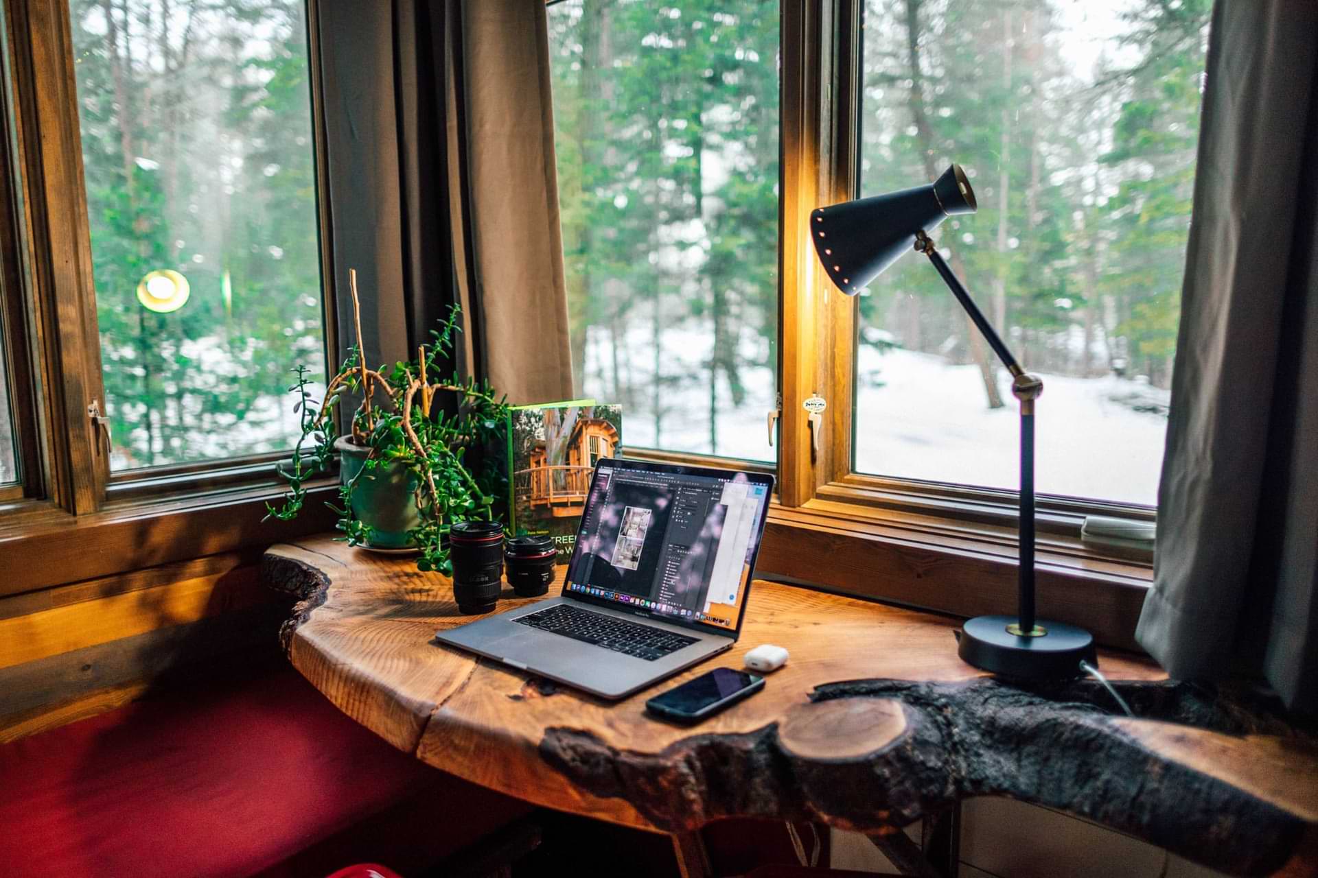 A Rustic Desk In A Cabin With A Laptop, Smartphone, Wireless Earbuds, Camera Lenses, Plants, And A Lit Lamp, Overlooking A Snowy Forest